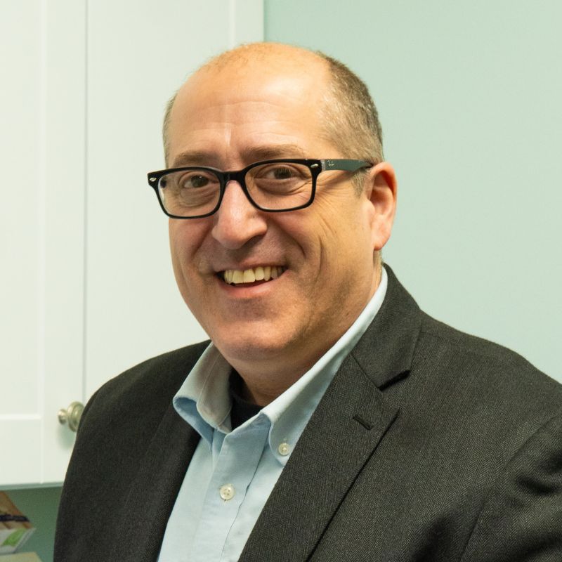 Dr. Stefan Fosco, audiologist, smiling in black glasses, a light blue shirt and a dark tweed jacket